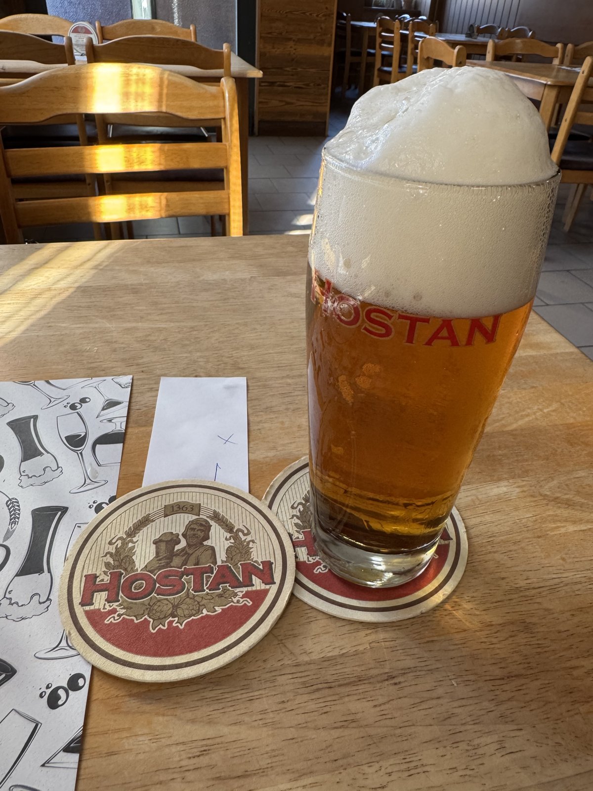 Půllitr piva Hostan na dřevěném stole s pivním táckem Hostan