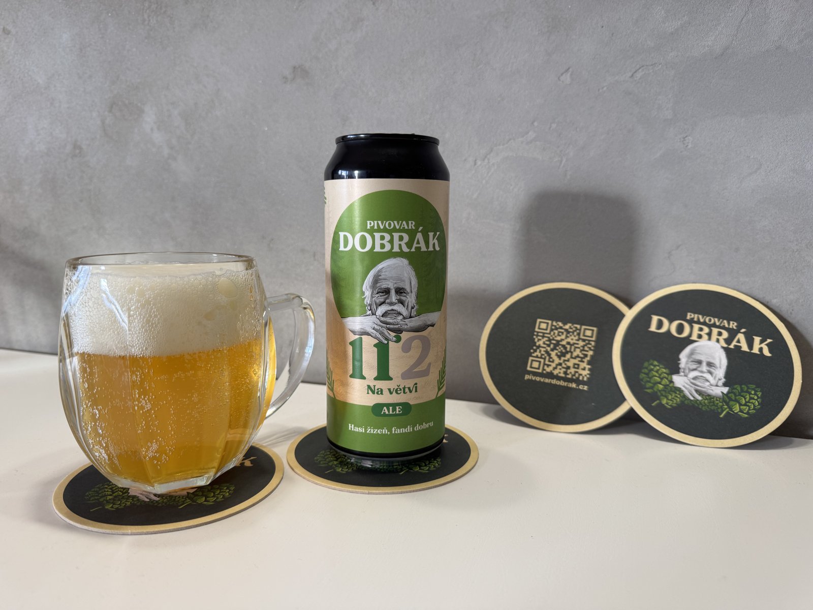 Pivovar Dobrák ALE se smrkovými výhonky v plechovce s načepovaným pivem a pivními podtácky