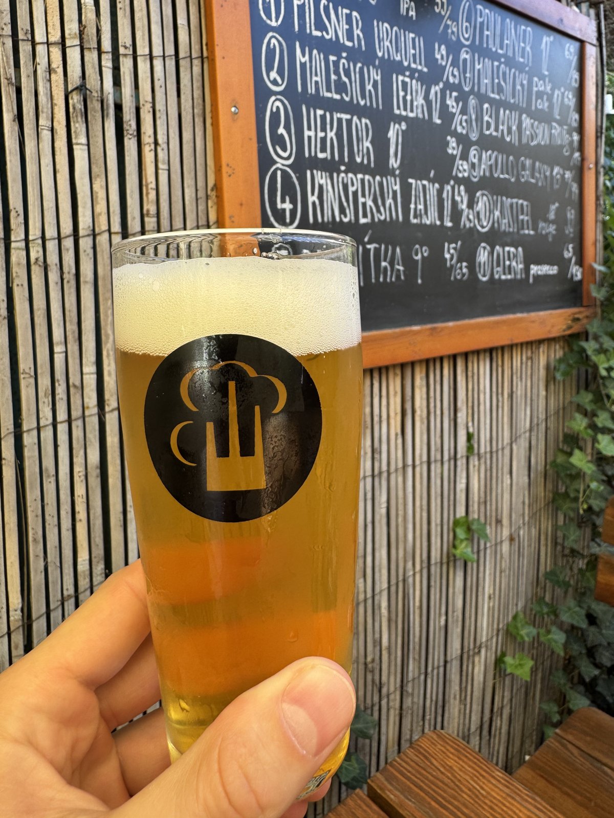 Malešický Pale Ale ve sklenici před tabulí s nabídkou piv